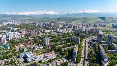 Aerial view of Bishkek city during spring, featuring lush greenery, urban buildings, and snowy mountains in the background under a clear blue sky