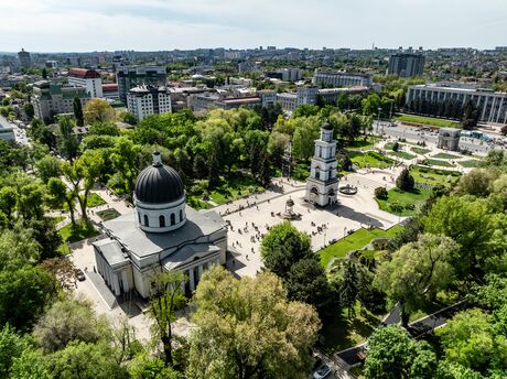 Aerial view of a church with a dome and separate bell tower in a park with many trees and surrounding cityscape