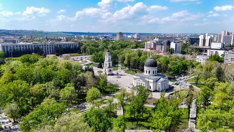 Aerial view of a city featuring a white church with a dome and a separate bell tower surrounded by many trees and other buildings in the background