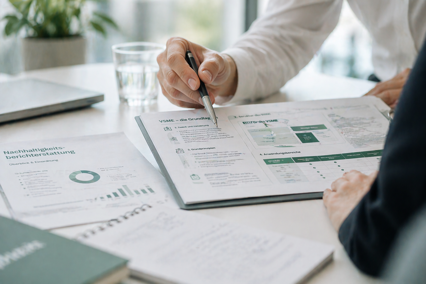 Zwei Personen besprechen Dokumente mit Diagrammen und Texten an einem Tisch mit Glas Wasser und Laptop im Hintergrund