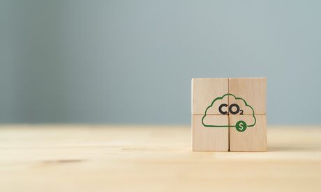 Two wooden blocks on a light wood surface with a green cloud containing 'CO₂' and a dollar sign against a gray background